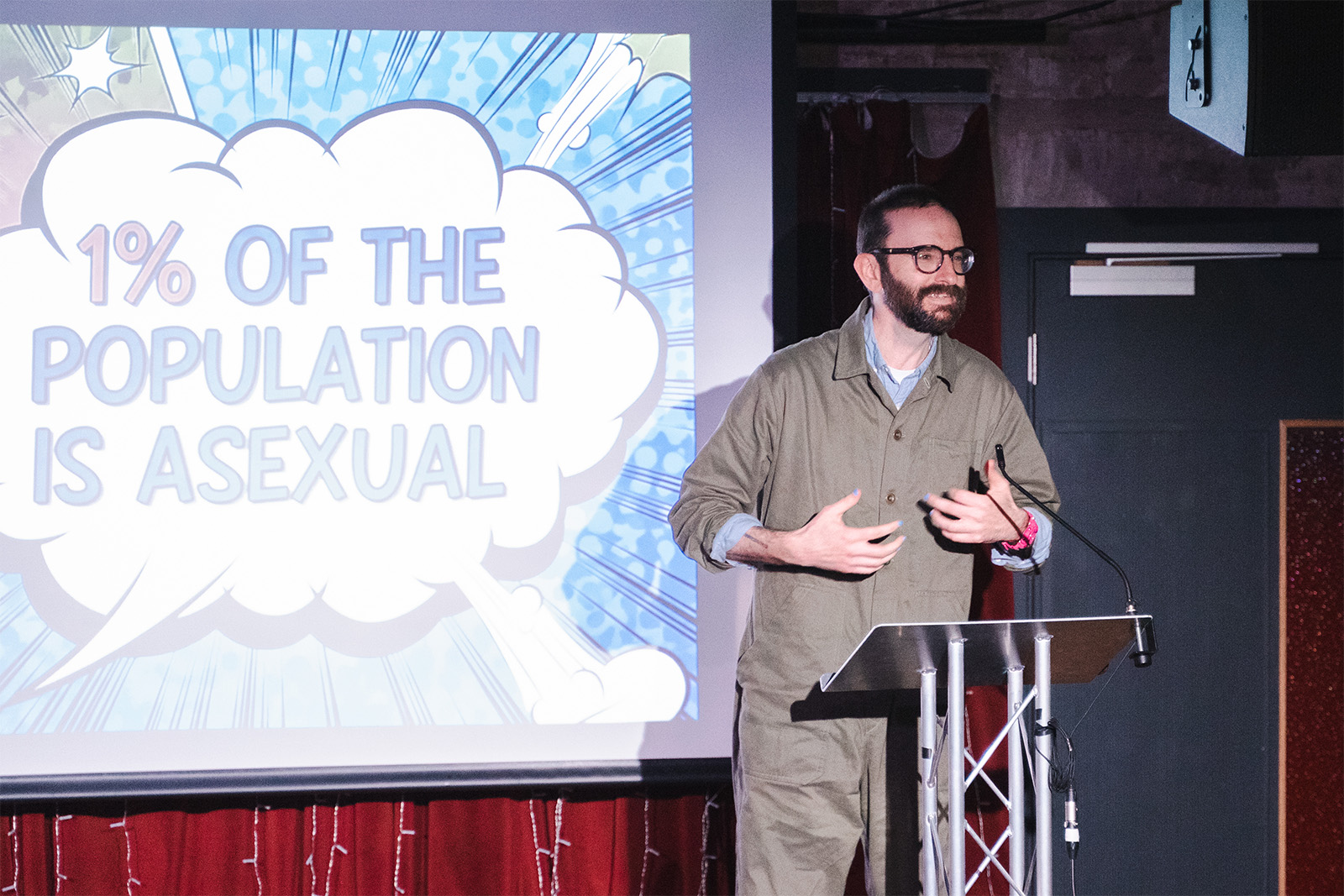 A presentation at the Queer Data Showcase. A person stands next to a large screen. The words '1% of the population is asexual' appear in large comic book-style lettering.