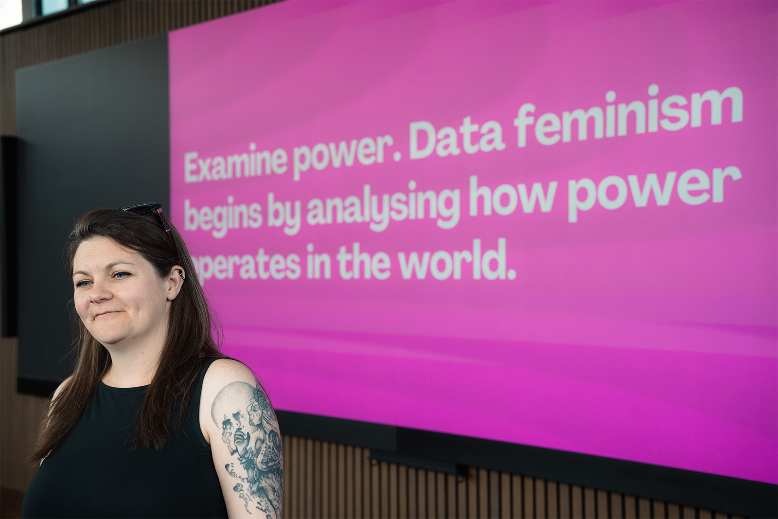 A speaker standing in front of a presentation screen. In a large bold font, the words on the screen read 'Examine power. Data feminism begins by analysing how power operates in the world.'