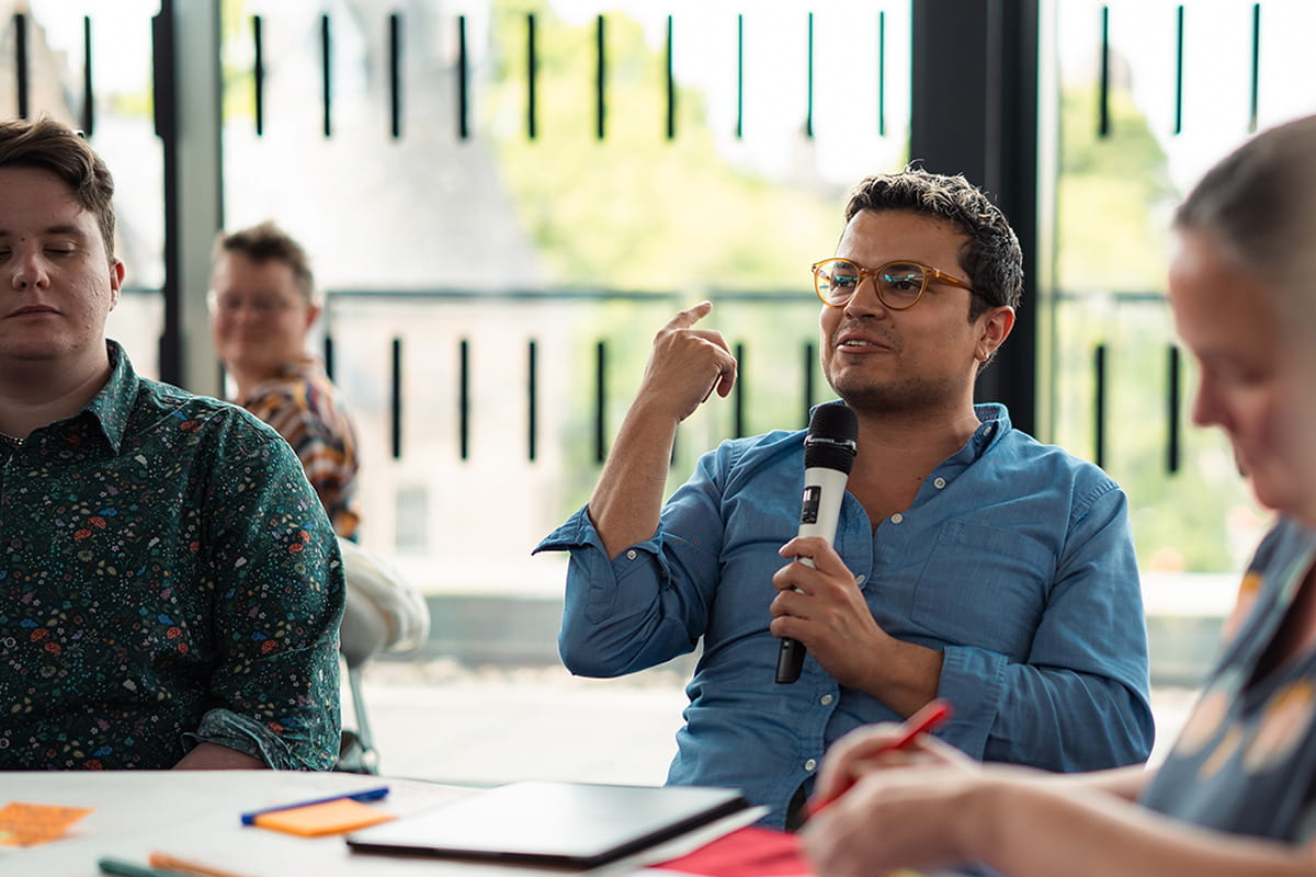 Four individuals seated around tables, with one person holding a microphone and addressing the audience